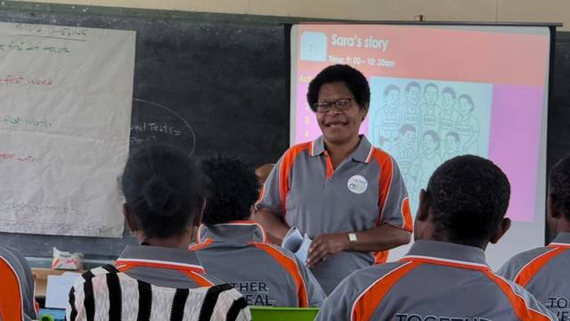 A facilitator in a programme shirt teaches a group, with a projected slide behind her.