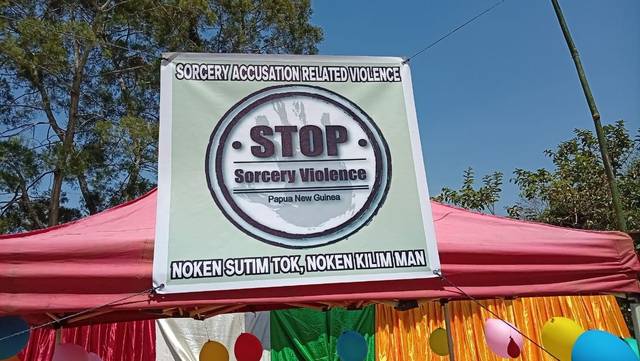 A hand-painted cloth banner reading Stop Sorcery Violence, Noken sutim tok, noken kilim man, hung outdoors at a graduation.