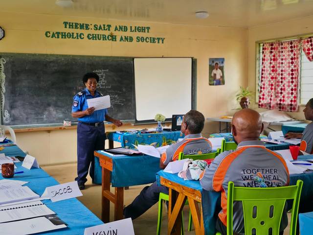 A police officer speaks to facilitators seated at tables in a church classroom.